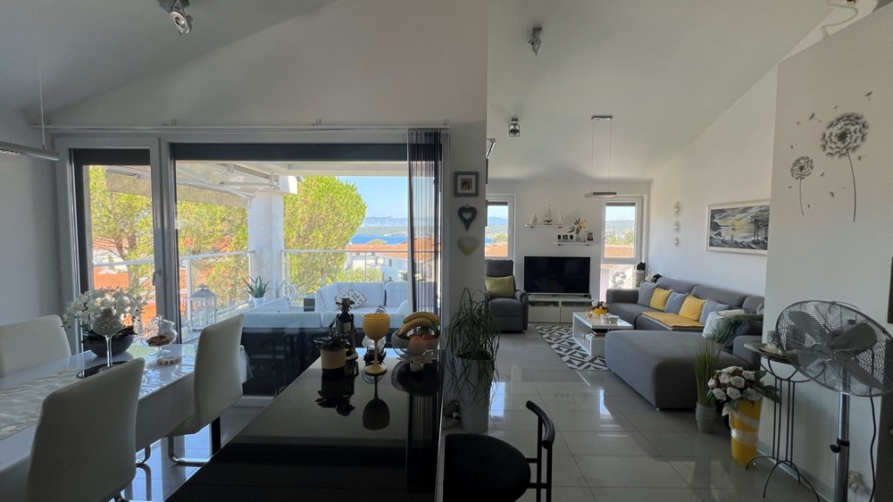 Ljust vardagsrum med grå soffa, matplats och stort fönster mot terrassen med havsutsikt.