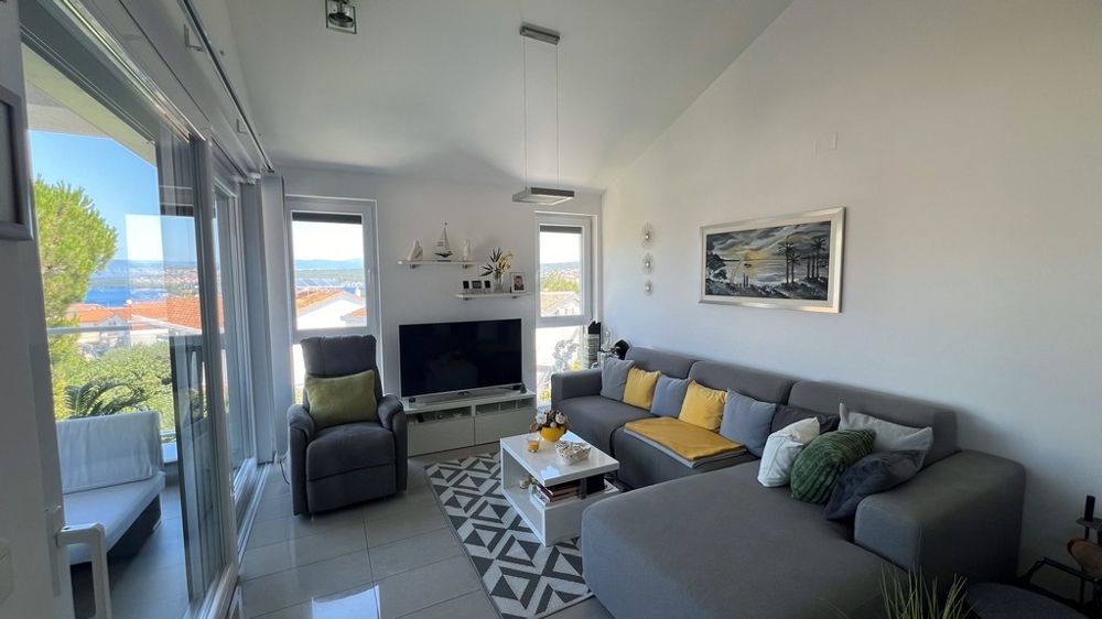 Modernt vardagsrum med stor TV, grå soffa och havsutsikt genom stora fönster.