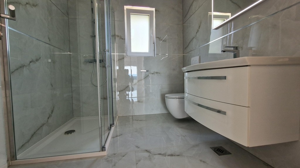 Modernt badrum med dusch, toalett och handfat, ljust kakel och fönster.