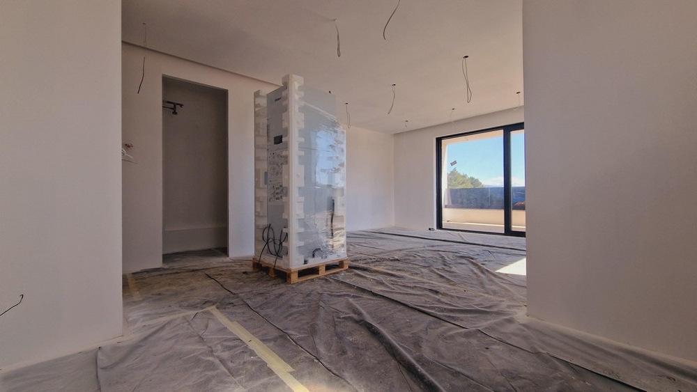 Stort vardagsrum i en modern nybyggnad, fortfarande under uppbyggnad, med fönsterfront och tillgång till terrassen.