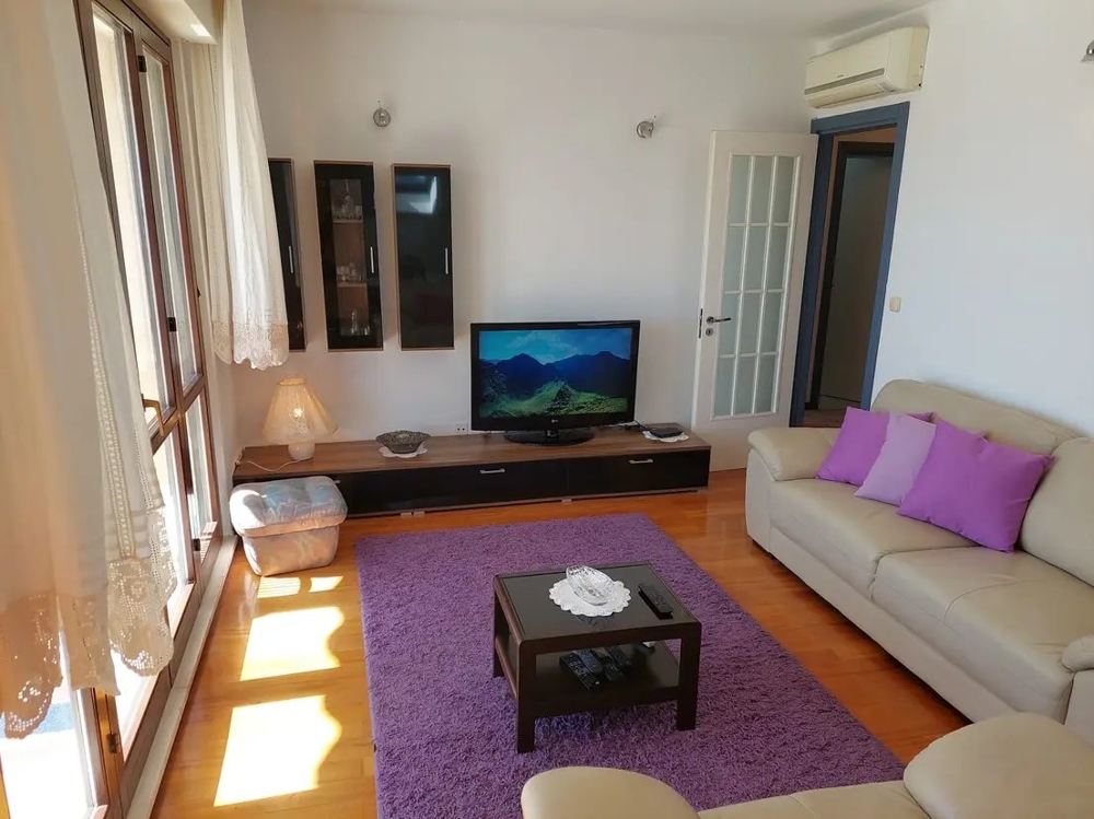 Vardagsrum med soffa, tv, lila kuddar och matta, stora fönster