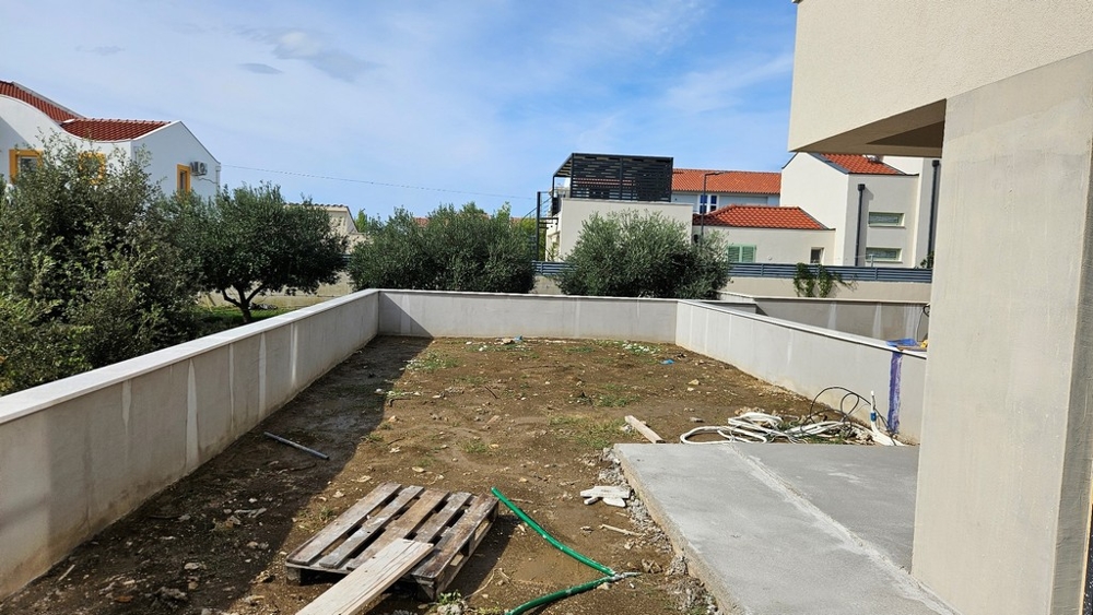 Uteområde med obanad mark, låg mur och några träd i bakgrunden.