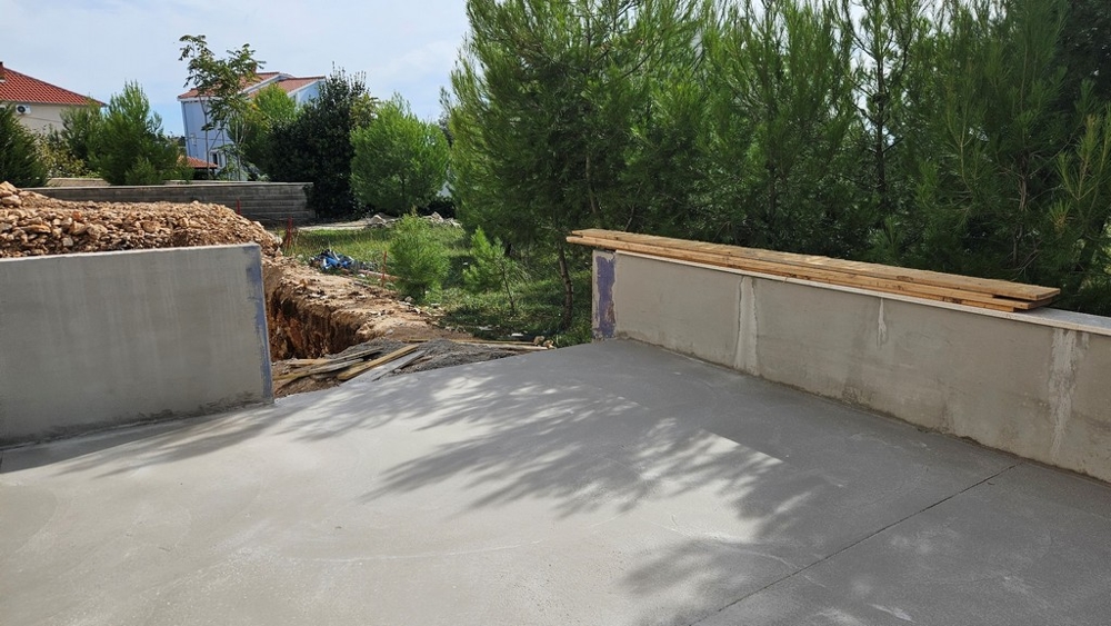 Delvis betong uteplats med byggplats och dike intill tallar.