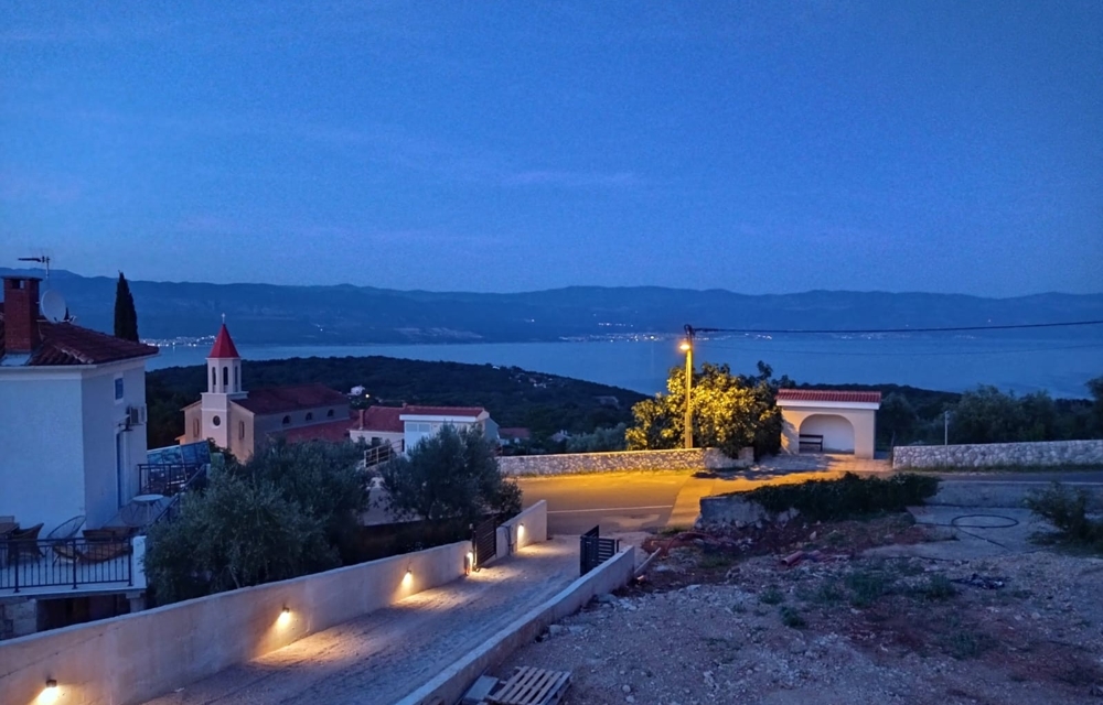 Utsikt från fastigheten i Kroatien över havet på natten, pool i förgrunden och upplyst by i bakgrunden.