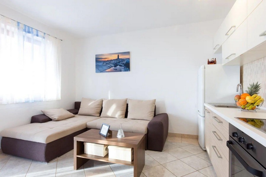 Mysigt vardagsrum med hörnsoffa, soffbord och utsikt över köket, inrett i mjuka färger och en bild på havet.