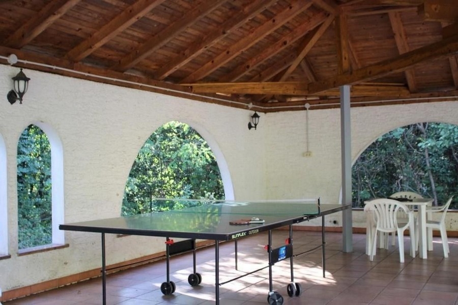 En täckt uteplats med ett bordtennisbord i mitten, vita plaststolar och ett trätak. Väggarna har välvda fönsteröppningar som ser ut mot gröna omgivningar.