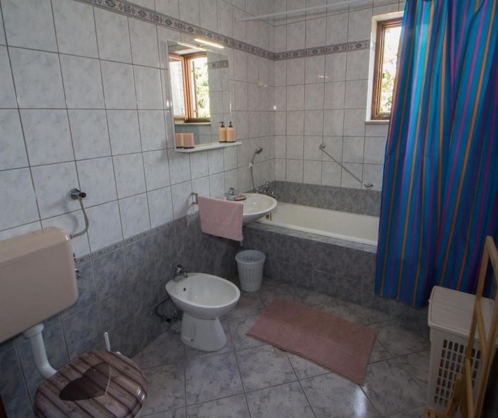 Ett badrum med vitt och grått kakel, ett badkar, ett handfat, en bidé, en toalett och ett blått duschdraperi. Små fönster släpper in naturligt ljus.