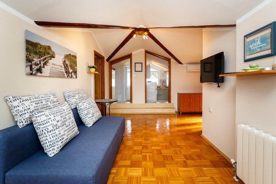 Vardagsrum med soffa, trägolv och takbjälkar leder till ett litet kök