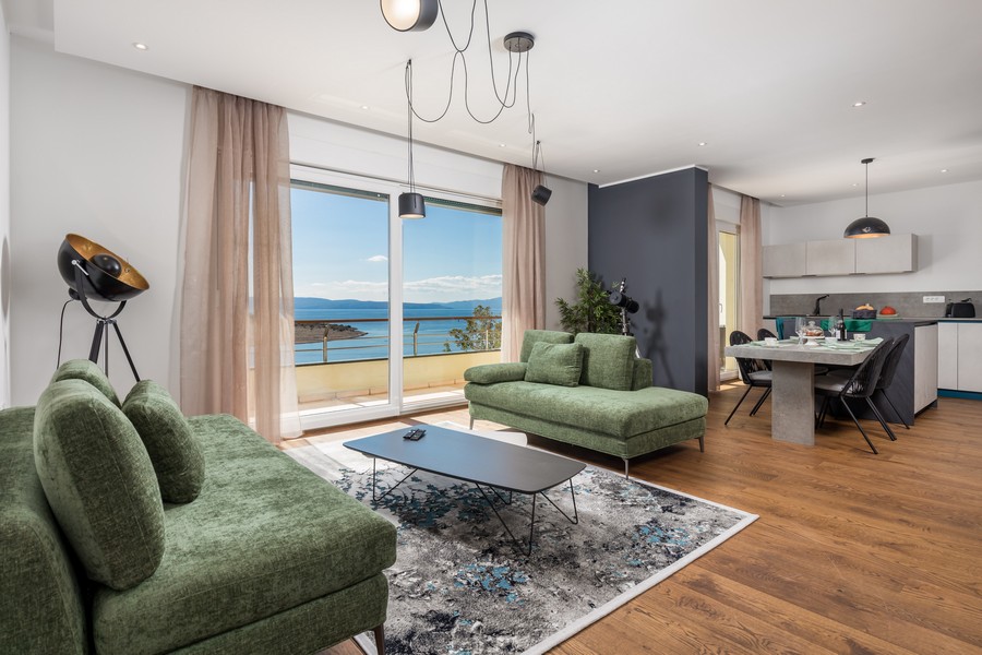 Vardagsrum med stora fönster, stilren inredning och havsutsikt, öppet kök i bakgrunden.