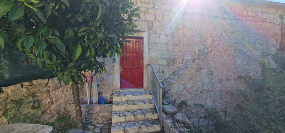 En liten uteplats med en stentrappa som leder till en röd trädörr. Husets vägg är gjord av natursten och solen skiner på himlen.