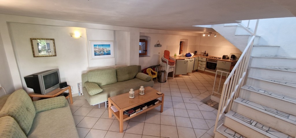 Ett mysigt vardagsrum i öppen planlösning med gröna soffor, ett soffbord, en TV och ett integrerat kök på baksidan. Ljusa kakelplattor täcker golvet.