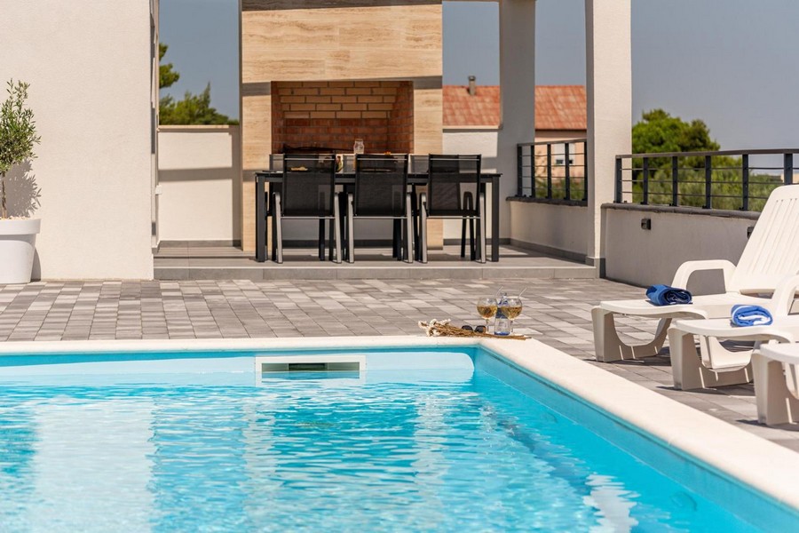 Uteplats med pool, solstolar och grillplats i bakgrunden.